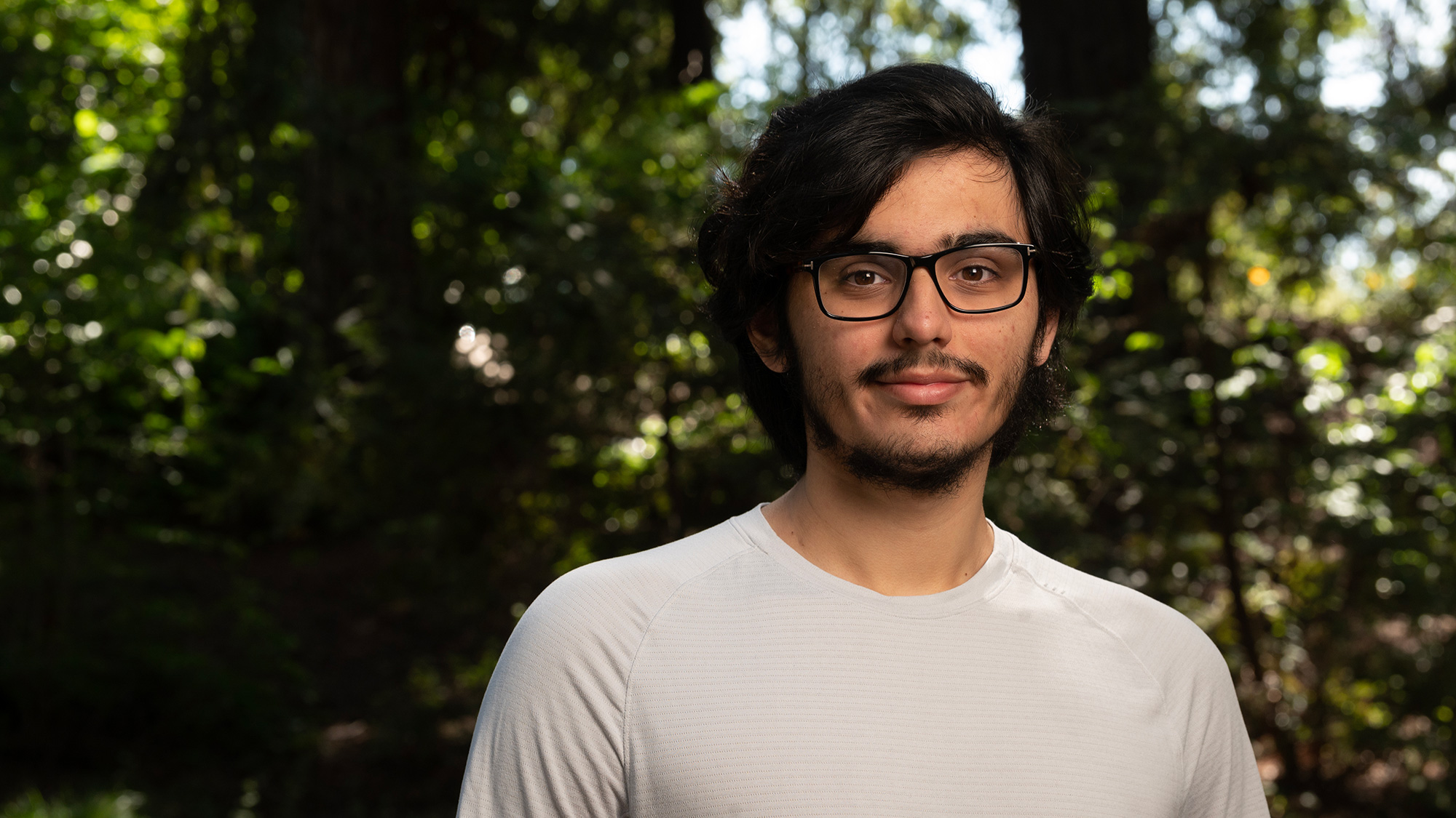 Casual outdoor portrait of a young man with glasses smiling against leafy background
