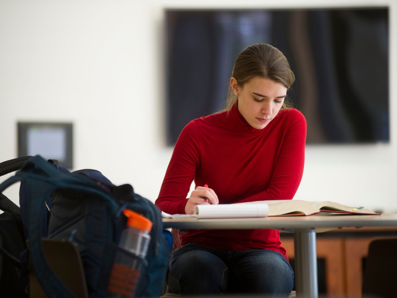 A student takes notes while reading from a textbook in a student lounge.