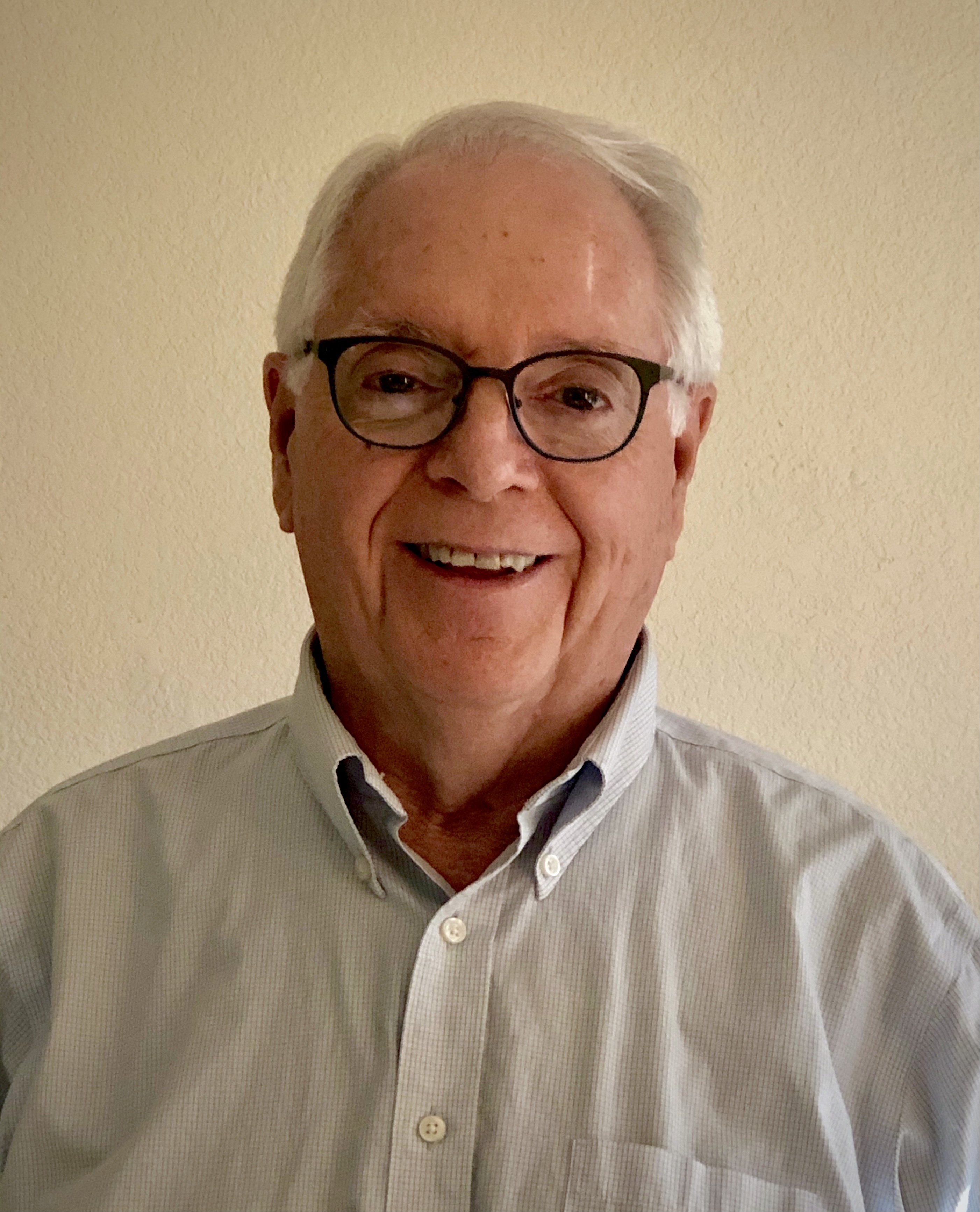 Distinguished Professor Michael Savageau is standing centered in front of the camera. He's smiling while wearing black rimmed glasses, a grey Oxford shirt and short white hair.
