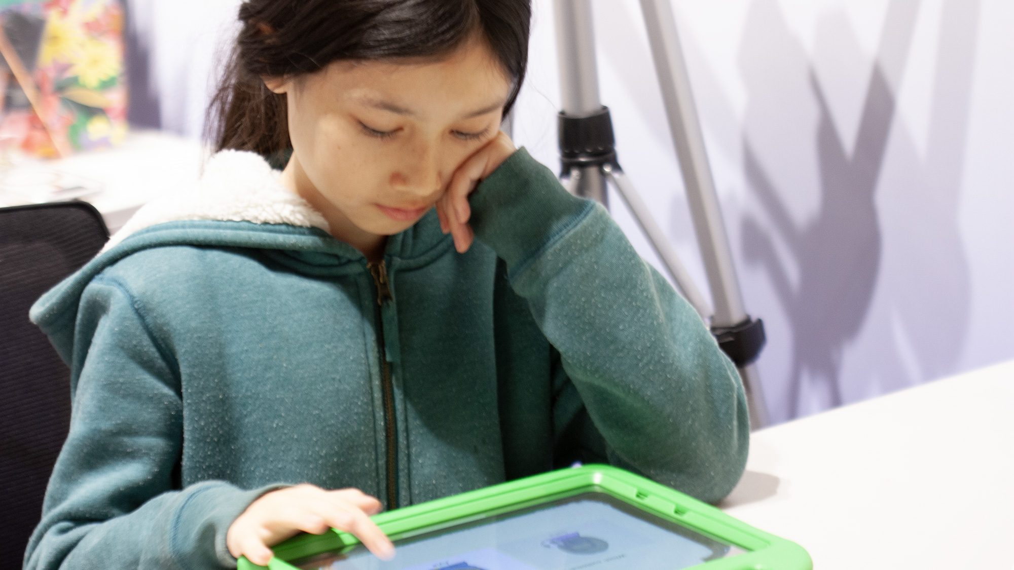Girl in green hoodie, resting head on hand while using green tablet