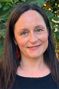 A woman with long dark hair smiles warmly, surrounded by green foliage.