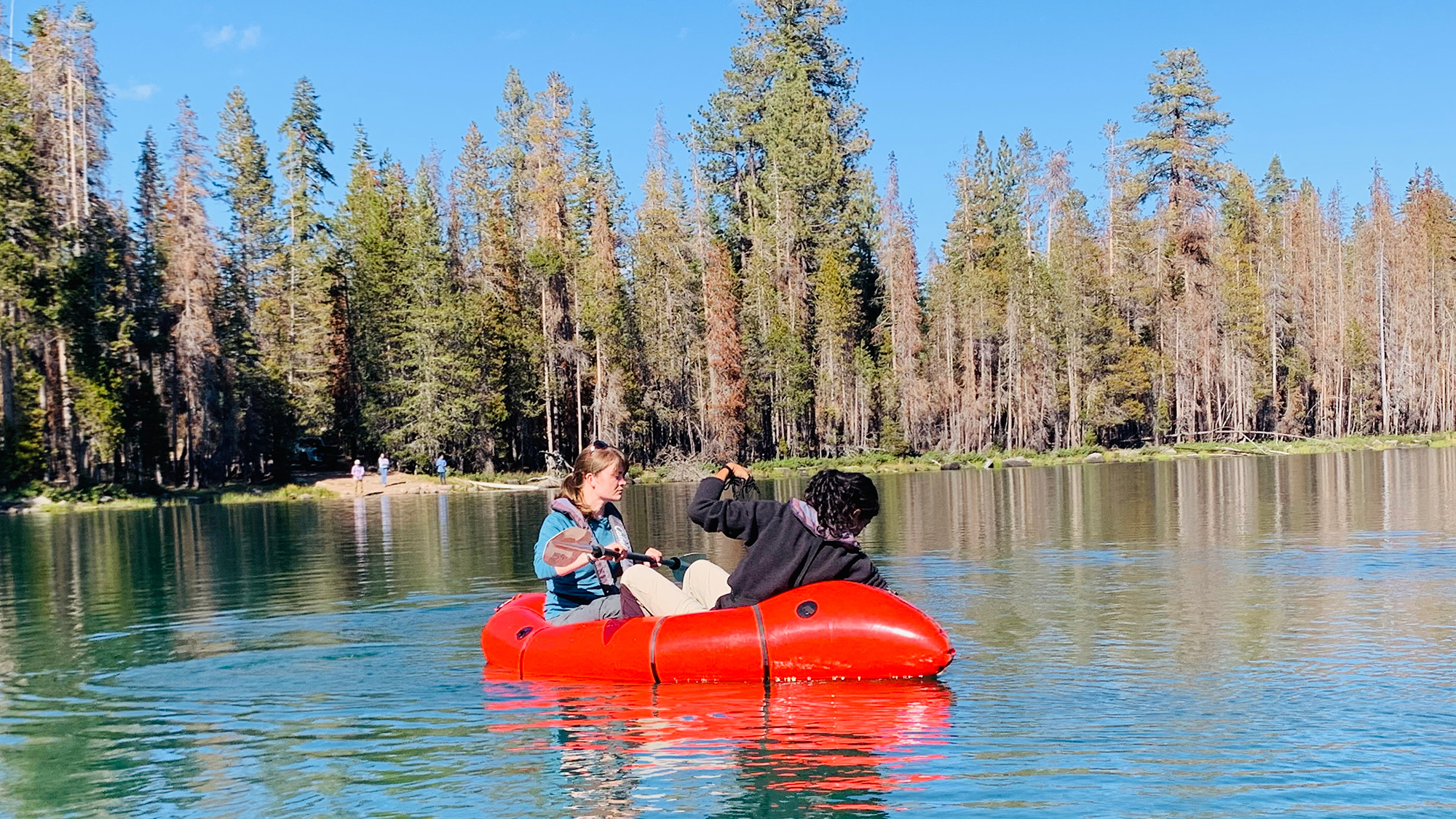 Two people in a red inflatable boat on a calm, sunny forest lake