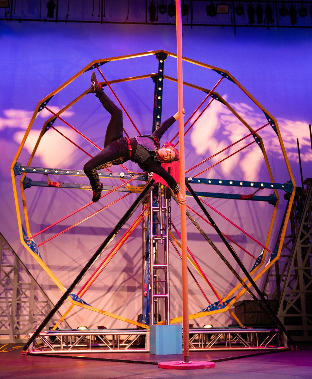 Acrobat upside-down inside large metal wheel on purple-lit stage