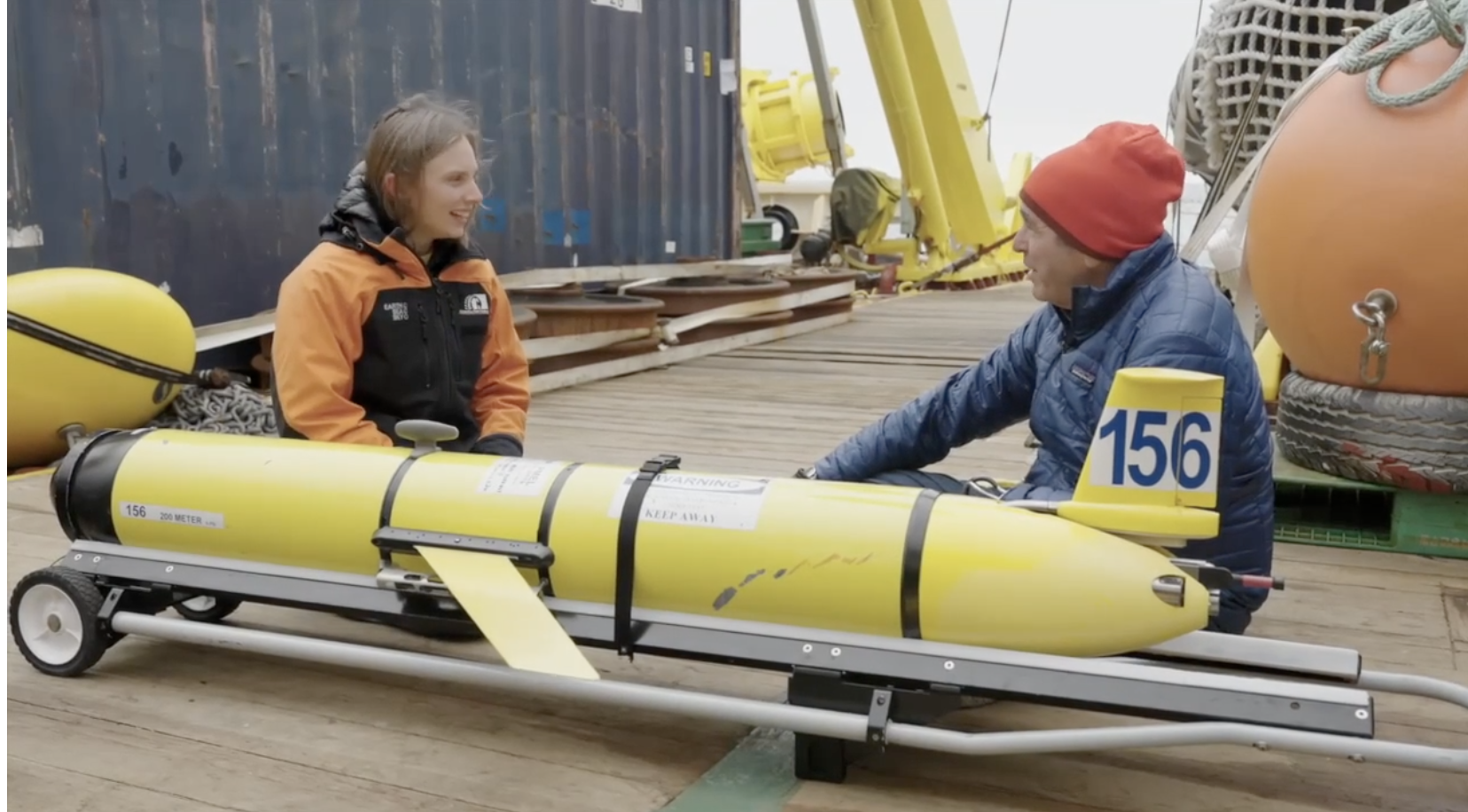 A woman and a man sit on a dock beside a yellow underwater drone and research equipment.