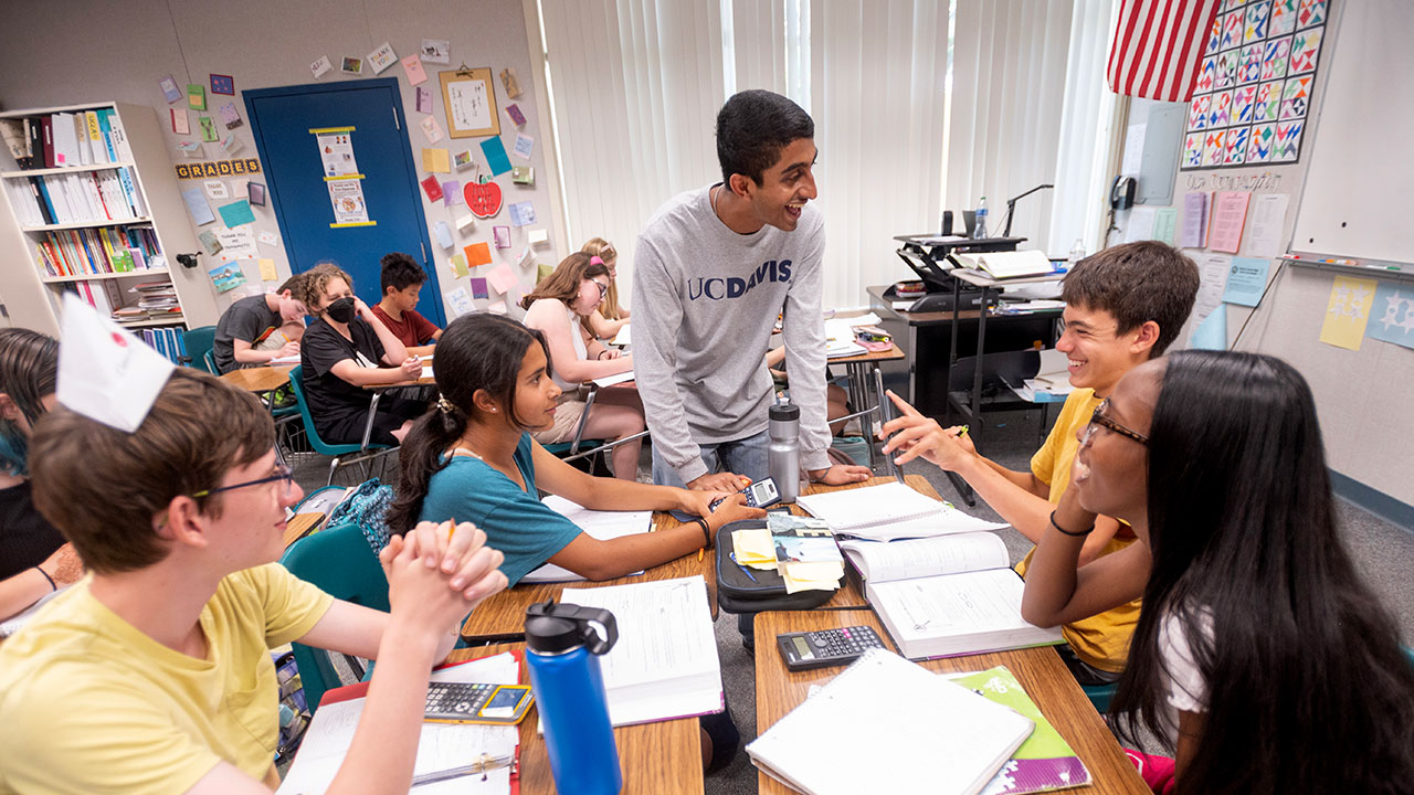 Standing, Neeraj Senthil talks with four students seated at their desks