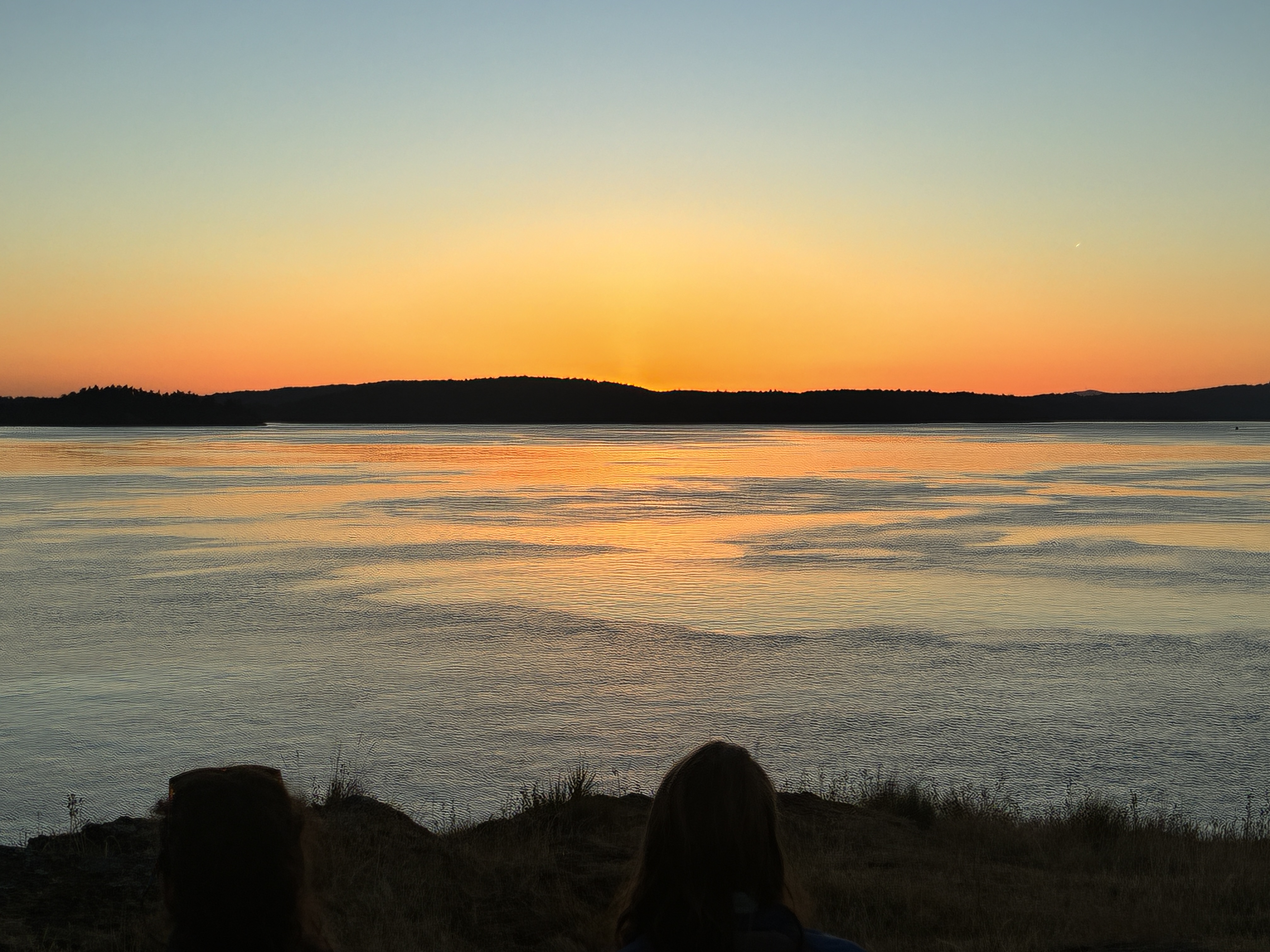 Orange sunset over calm water with two silhouetted people on a grassy bluff
