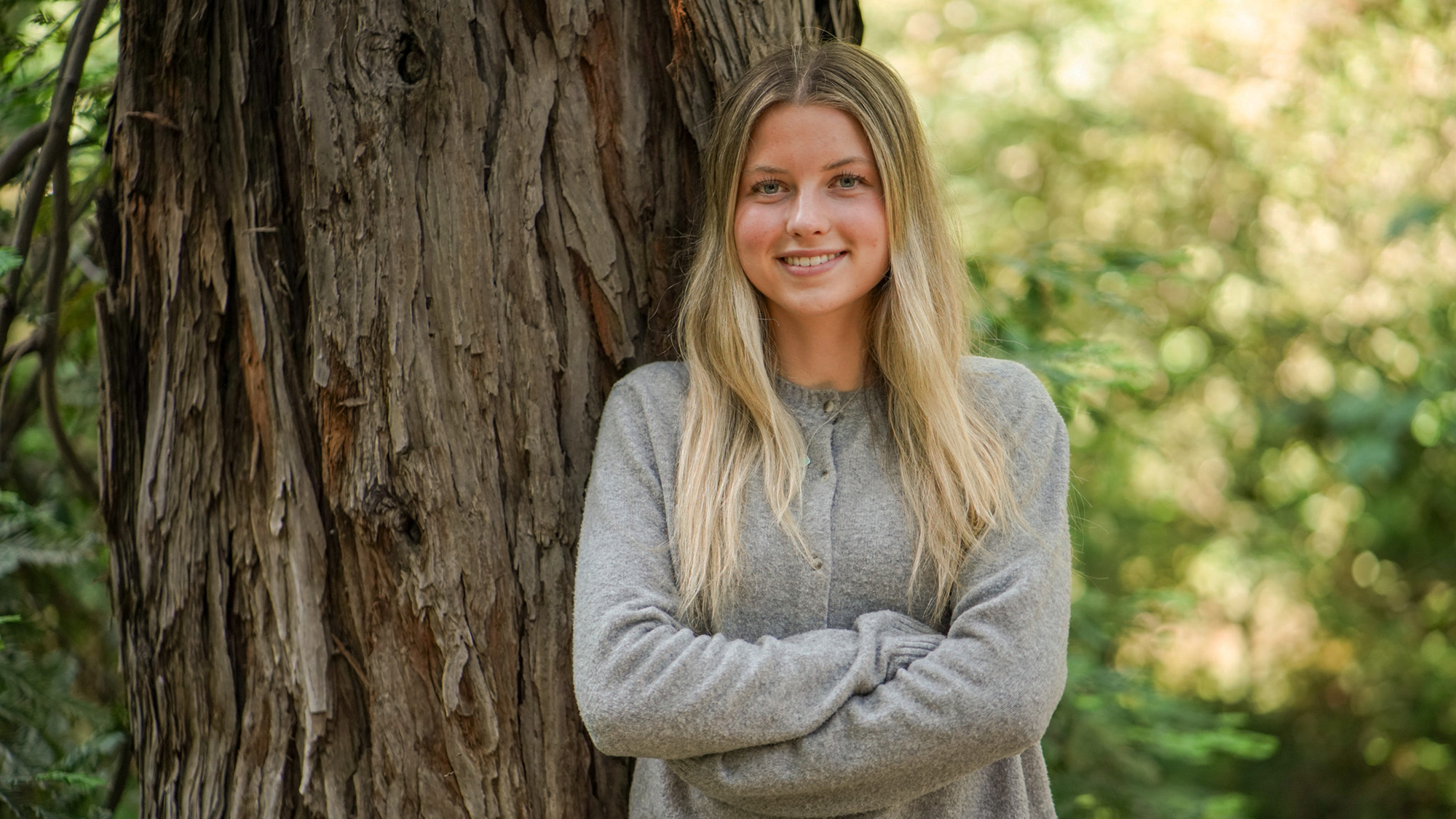 Smiling young woman with blonde hair, gray sweater, arms crossed by a tree outdoors