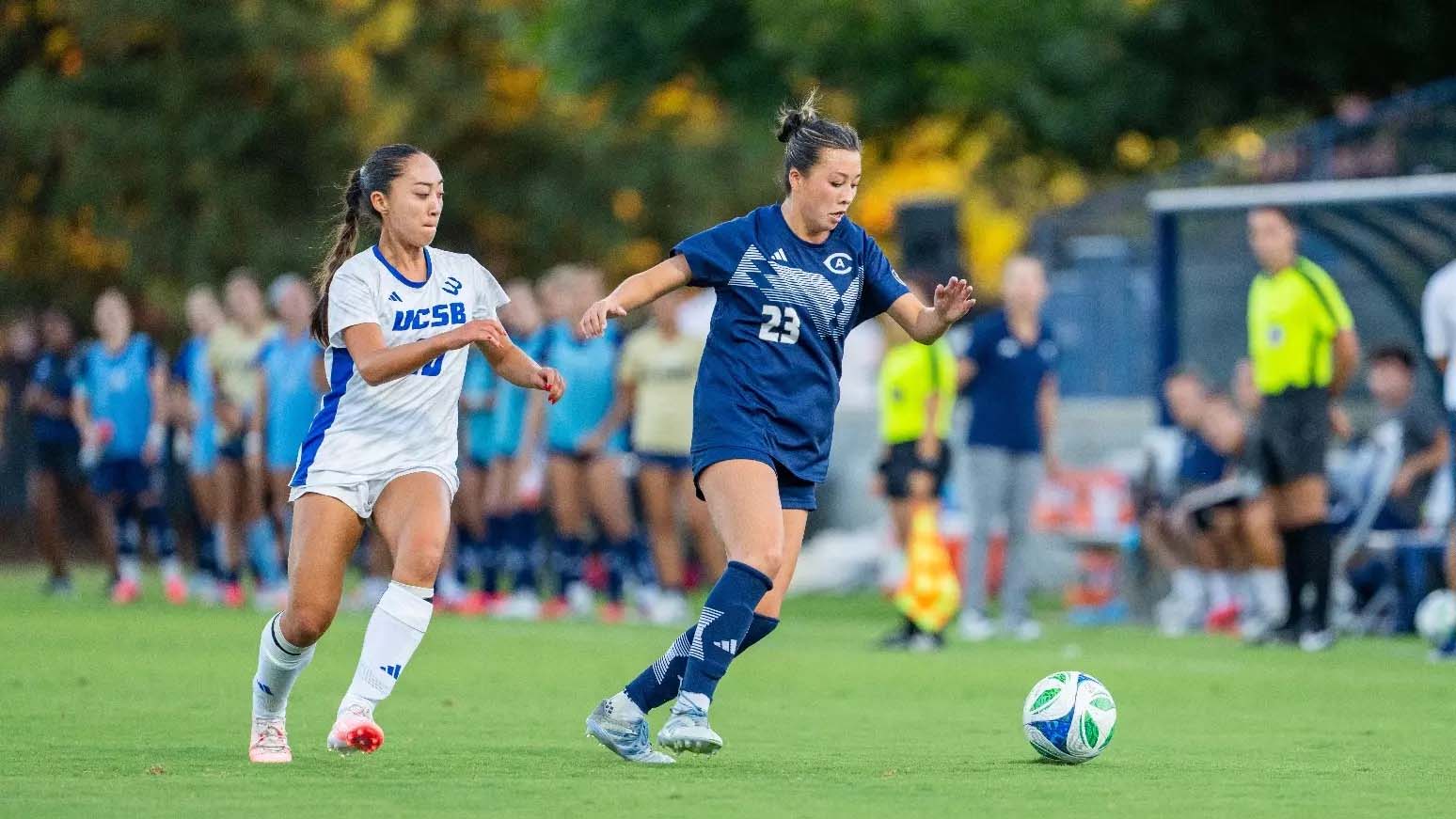 Two female soccer players — navy and white uniforms — sprinting toward ball on grass, blurred crowd