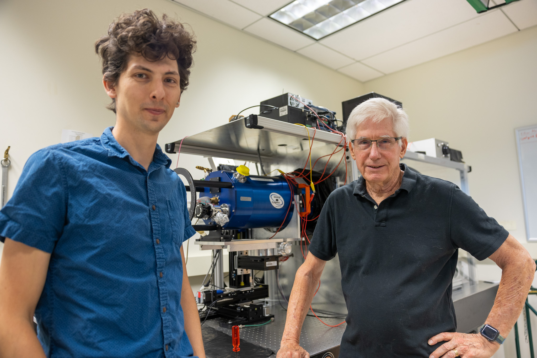 A young man with brown curly hair and a blue shirt and an older man with grey hair and a dark blue shirt look towards the camera in an indoor lab. Behind them is an apparatus with wires and cylinders.