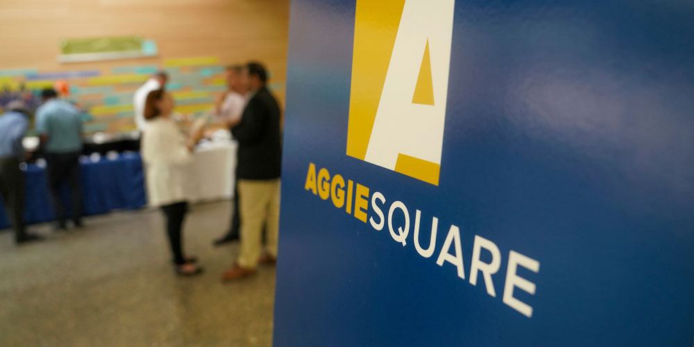 A sign with the Aggie Square logo and a few people talking in the background