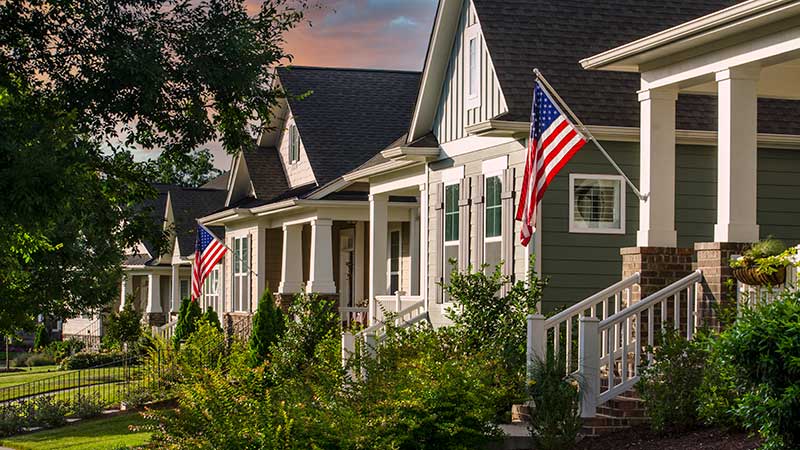 Suburban street scene with modest Craftsman style homes, American flags and green lawns.