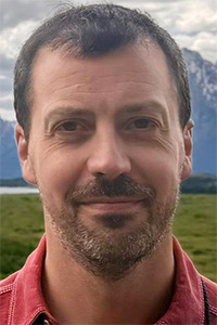 Man with short dark hair and stubble smiling against mountain and cloudy sky backdrop