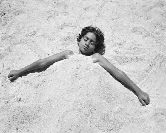Child buried up to neck in white sand, head and outstretched arms, relaxed face