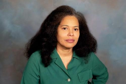 Woman with light brown skin, dark shoulder length hair wearing medium green top against neutral medium gray background.