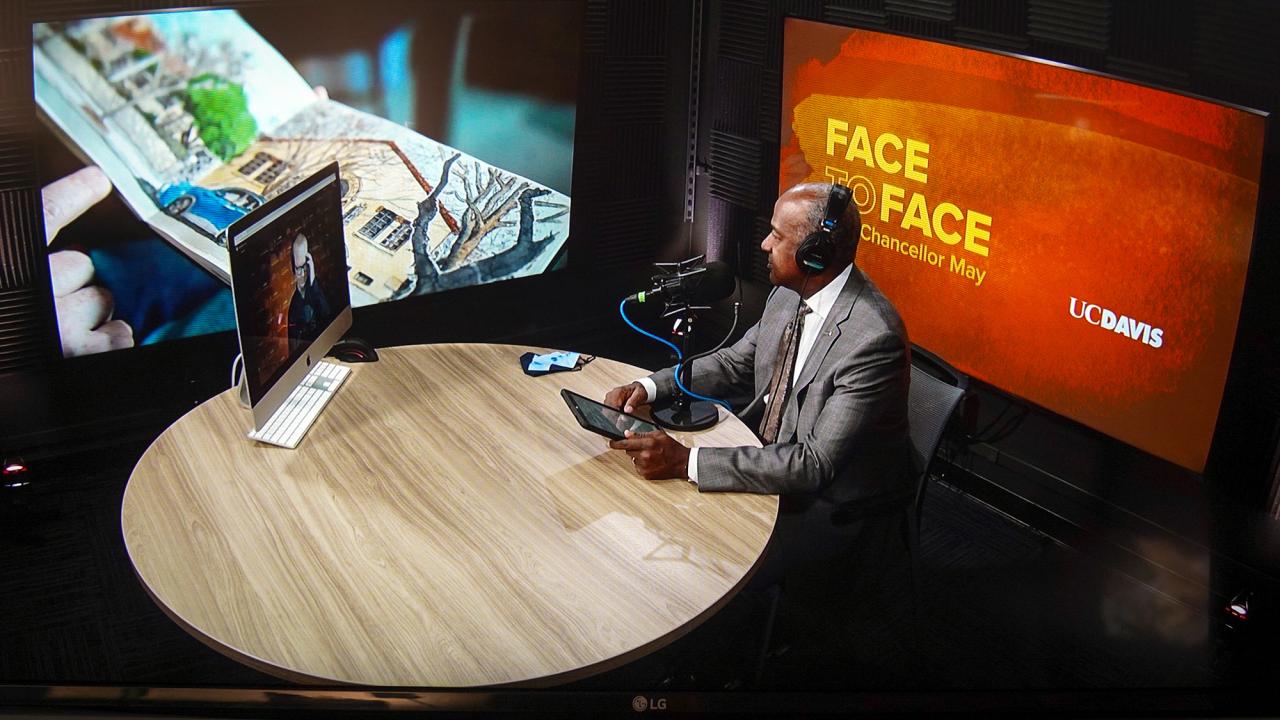 Chancellor Gary May sits at a round table with a microphone and speaks to Pete Scully through a video call on his computer.