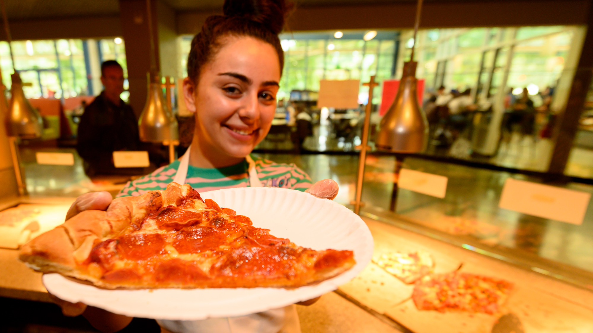 Counter person holds out a slice of pizza on a paper plate