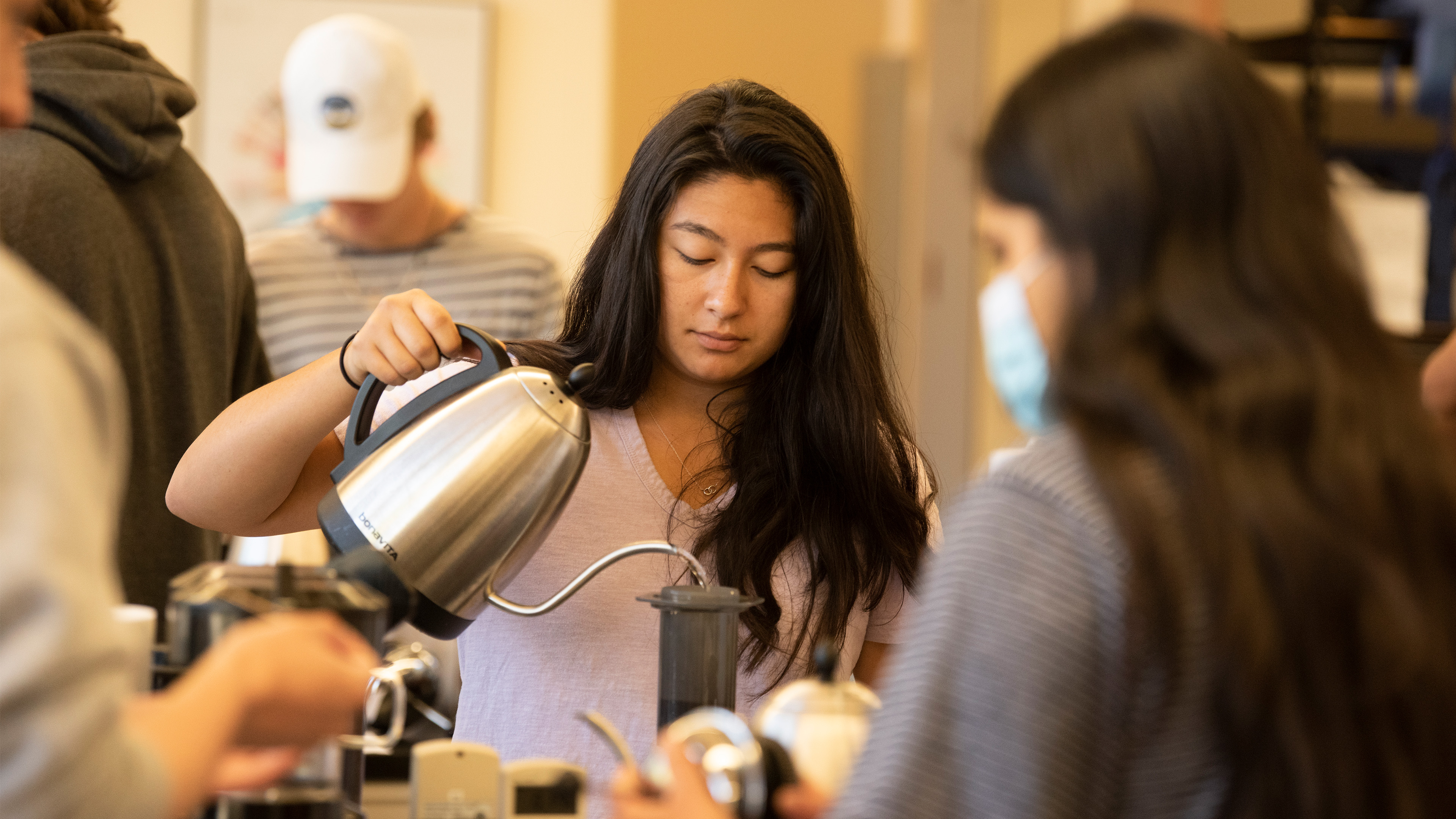 A student with long, dark hair pours boiling water from a stainless-steel kettle over coffee grounds in a dark cylinder.