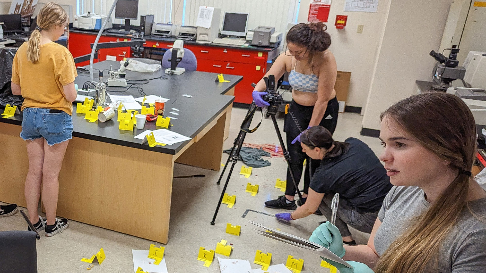 Students placing many yellow crime scene tags across a science lab bench while filming with a tripod.