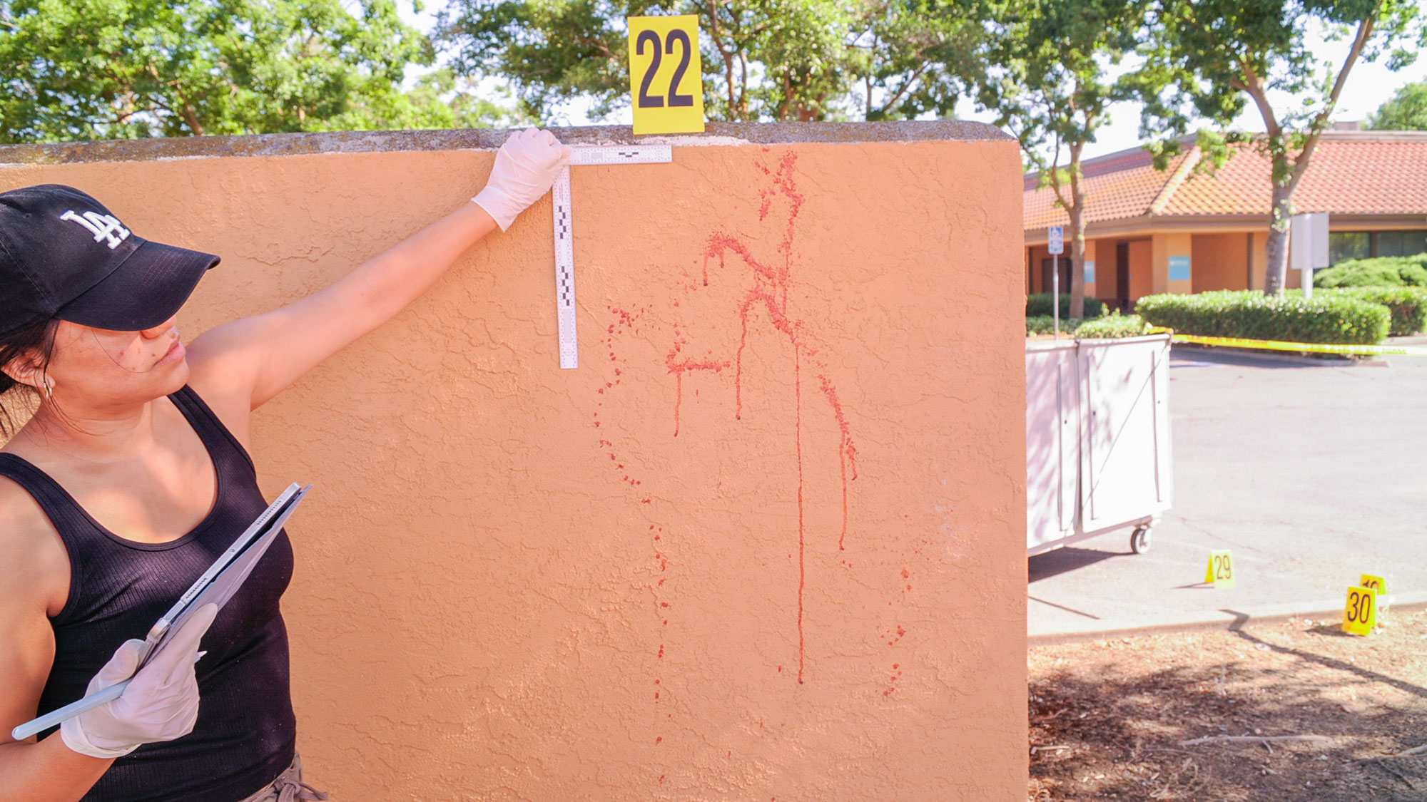 Woman analyzes red splatters on a wall with a yellow crime scene tag labeled 22