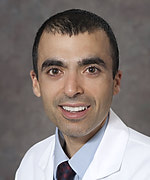 a man in a lab coat, shirt and tie, with short black hair, smiles towards the camera
