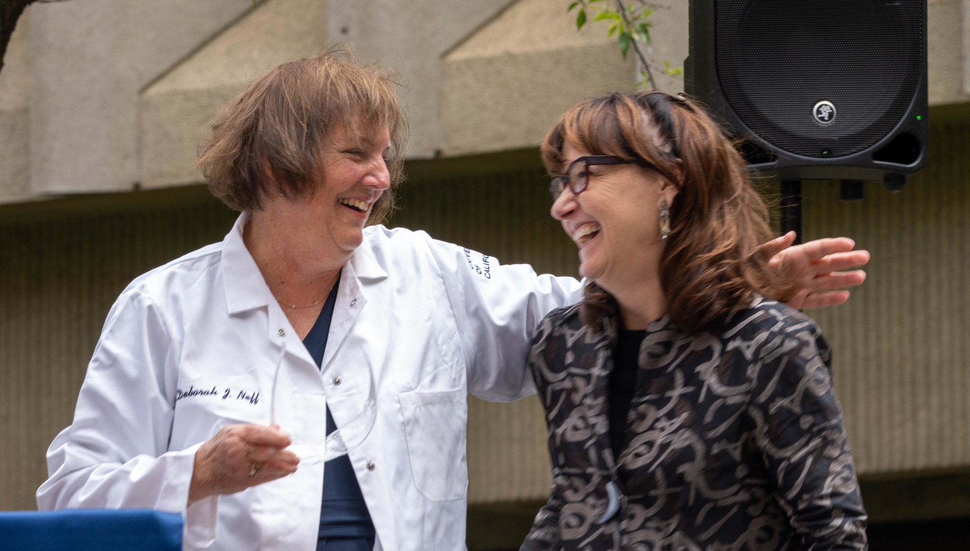 Woman in lab coat throws arm around woman in gray-black suit