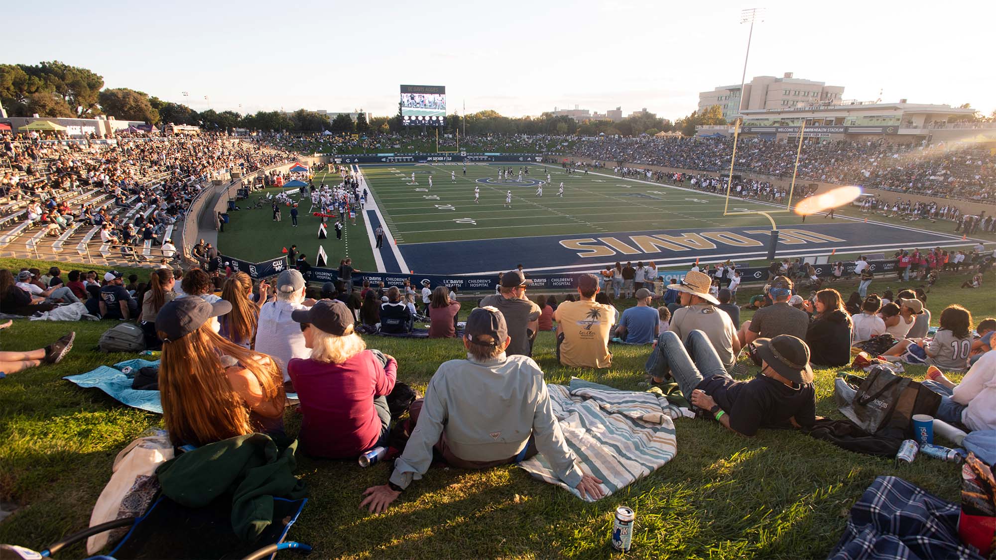 People on grassy hill watching a packed college football stadium at sunset