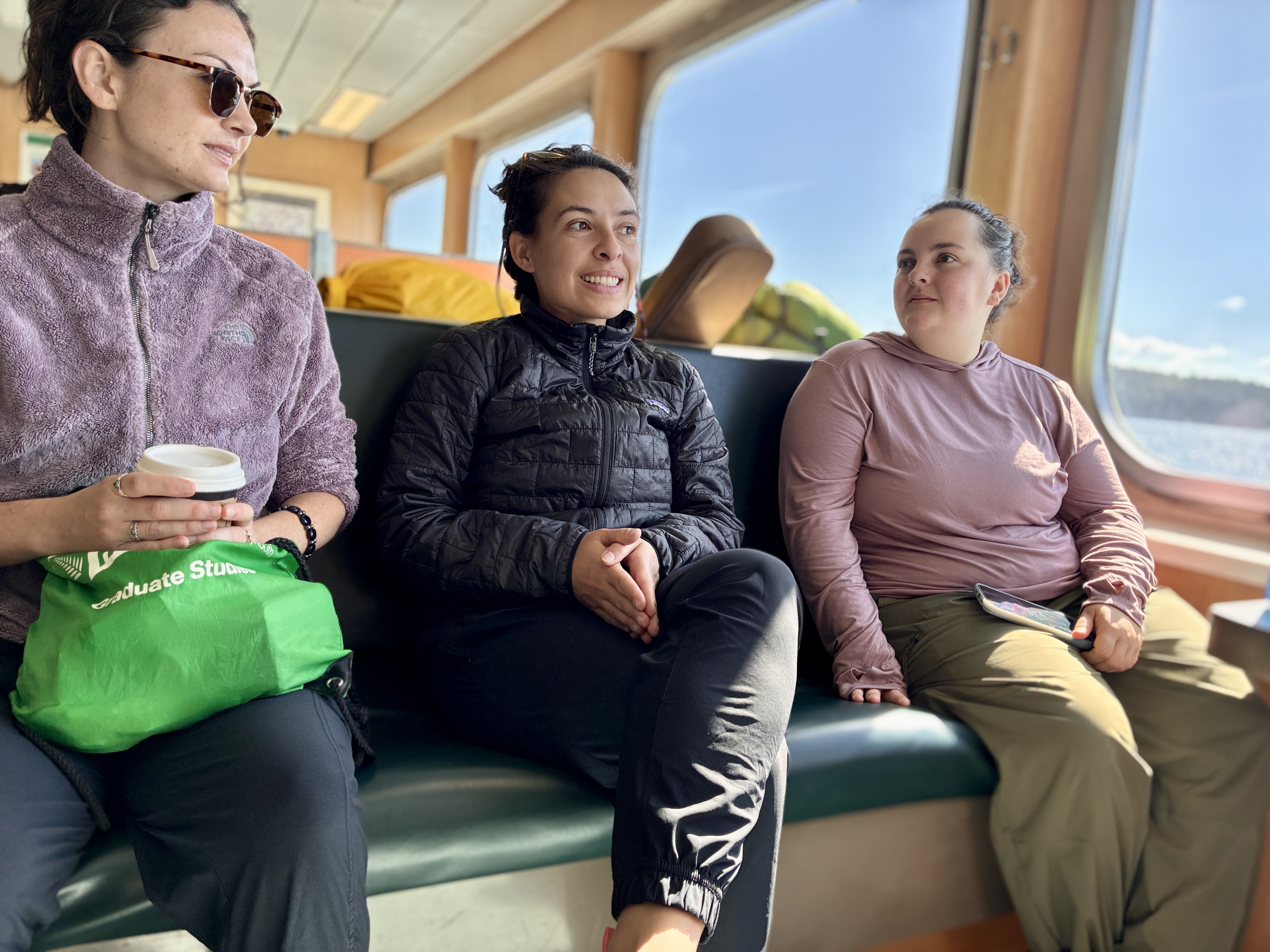 Three passengers sitting on a sunlit boat bench by large windows, chatting