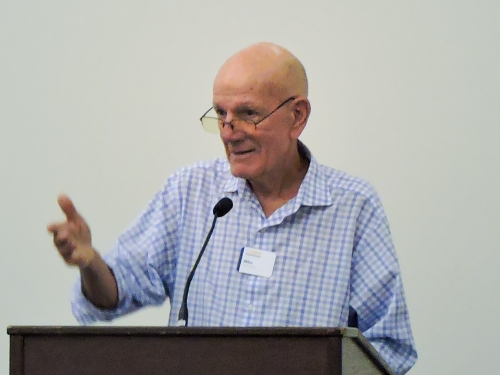 Elderly man in glasses smiling and speaking at a podium with a microphone