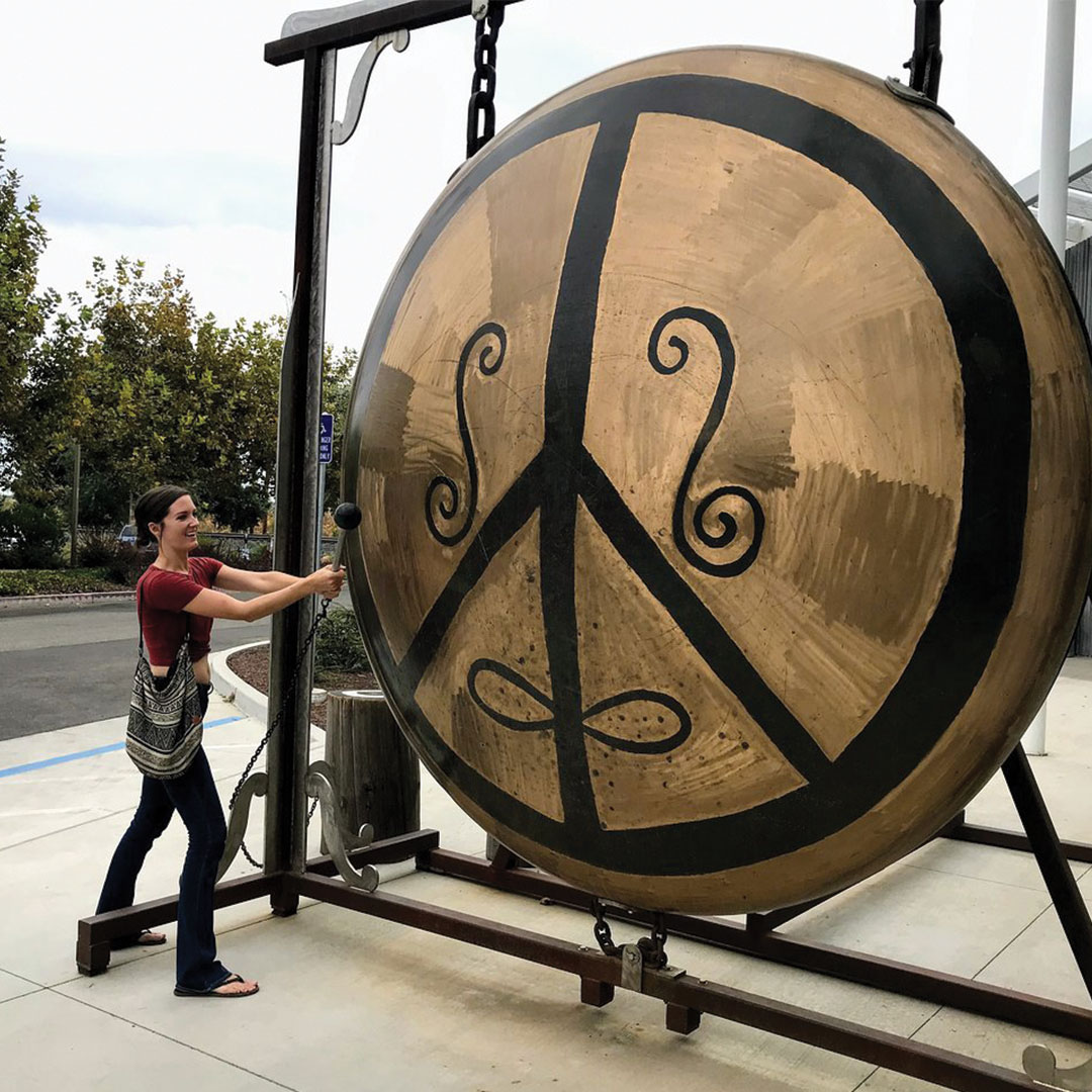 Person striking a giant bronze gong with a peace-symbol design outdoors