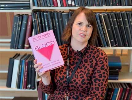 Woman in library holds up copy of book: Blood: The Science, Medicine, and Mythology of Menstruation, by Dr. Jen Gunter