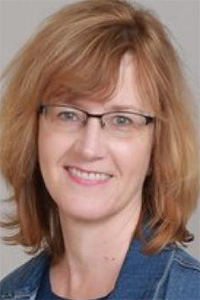 Smiling woman with glasses, shoulder-length light brown hair, denim jacket, gray background.