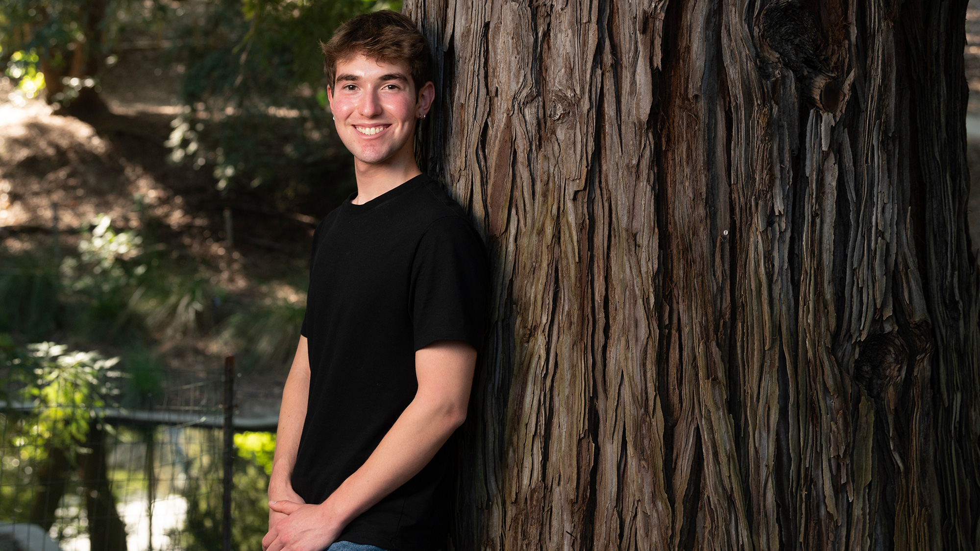 Young man in black tee leaning on large tree trunk, sunlit outdoor portrait photograph
