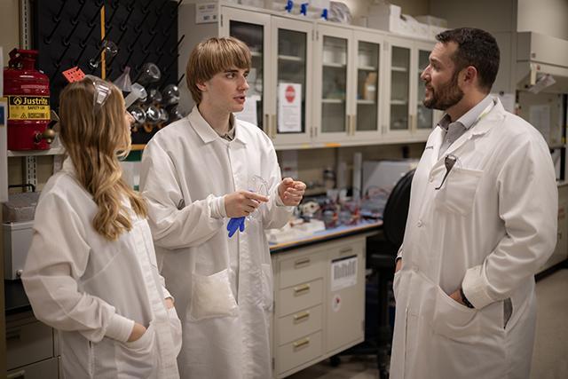 Three people in lab coats discussing in a laboratory setting with equipment and cabinets.
