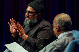 Two men, seated, on stage, in discussion