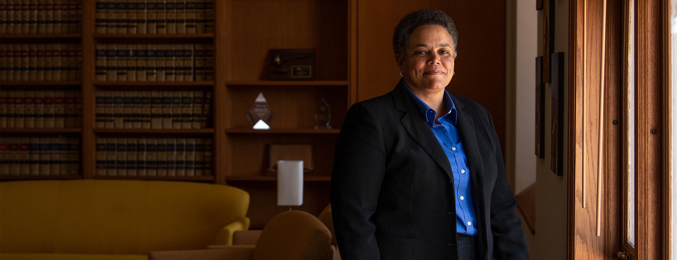 Judge Kelli Evans, wearing a bright blue button-up and black jacket, stands in the UC Davis law school building for a photo. Light flows in through a window, and a mustard couch and shelves of old law books are in the background.