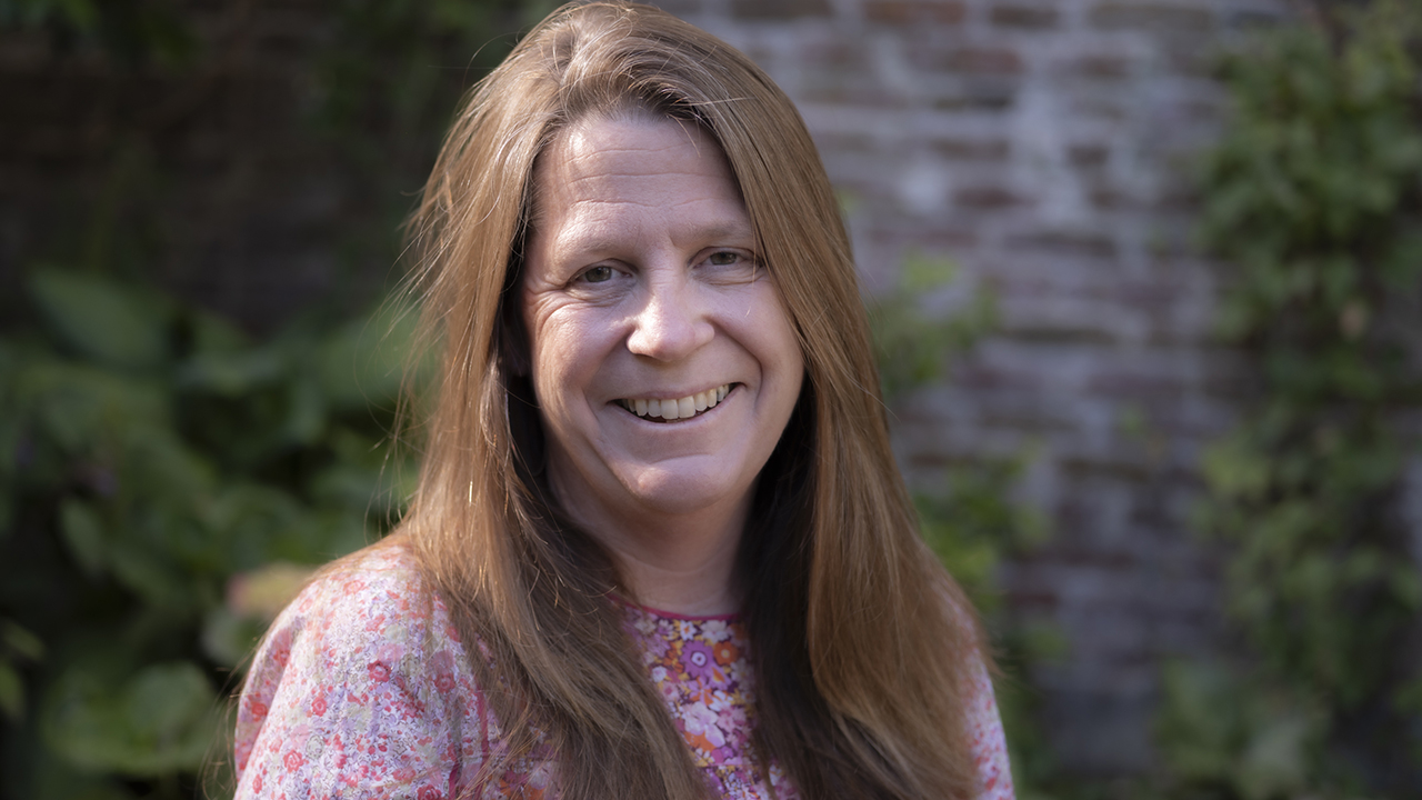 Outdoor headshot of a smiling woman with long hair against a brick wall and plants