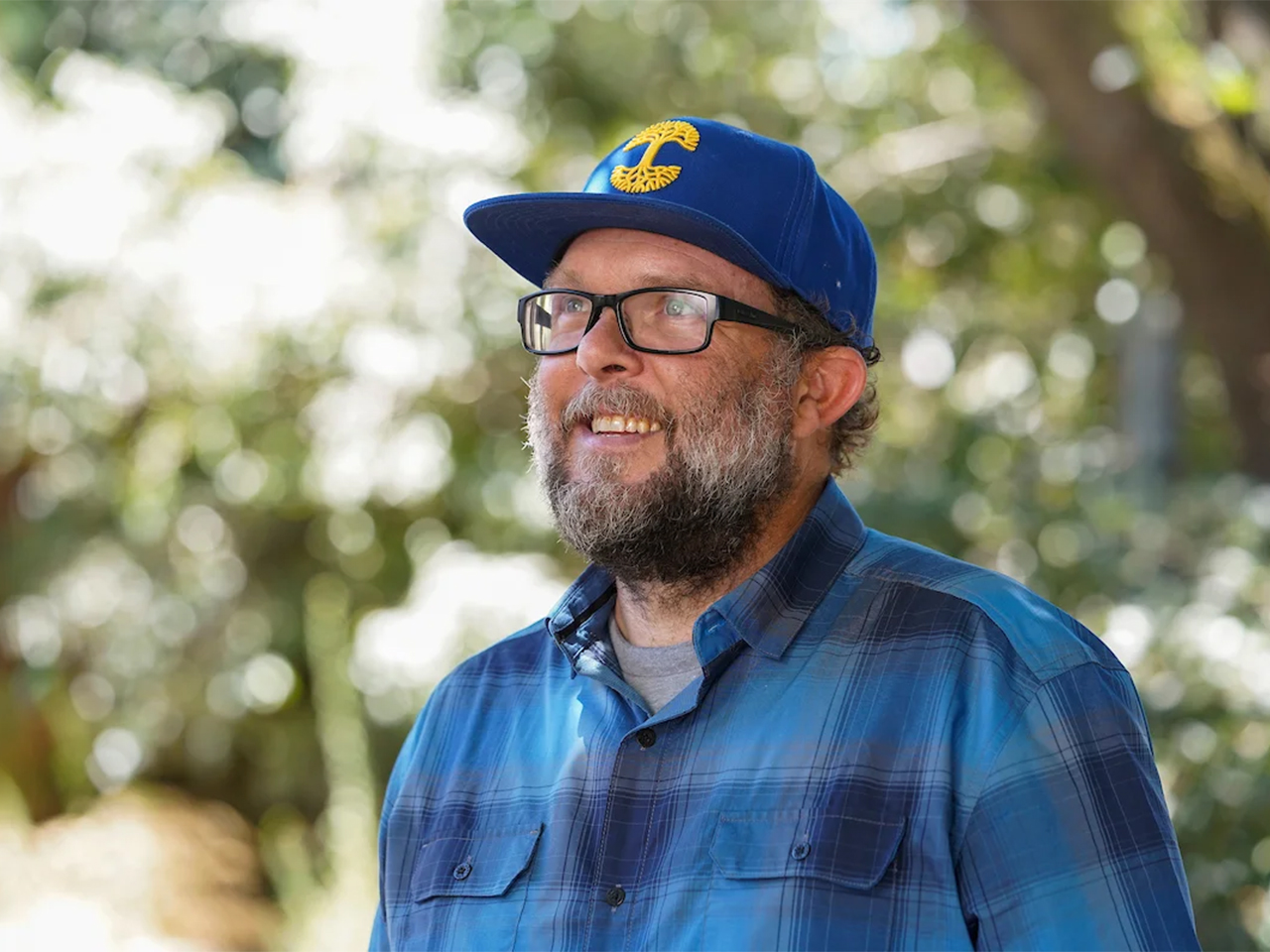 Keith Bein in a blue plaid shirt, glasses and blue baseball cap smiles, looking in the distance with trees in the background
