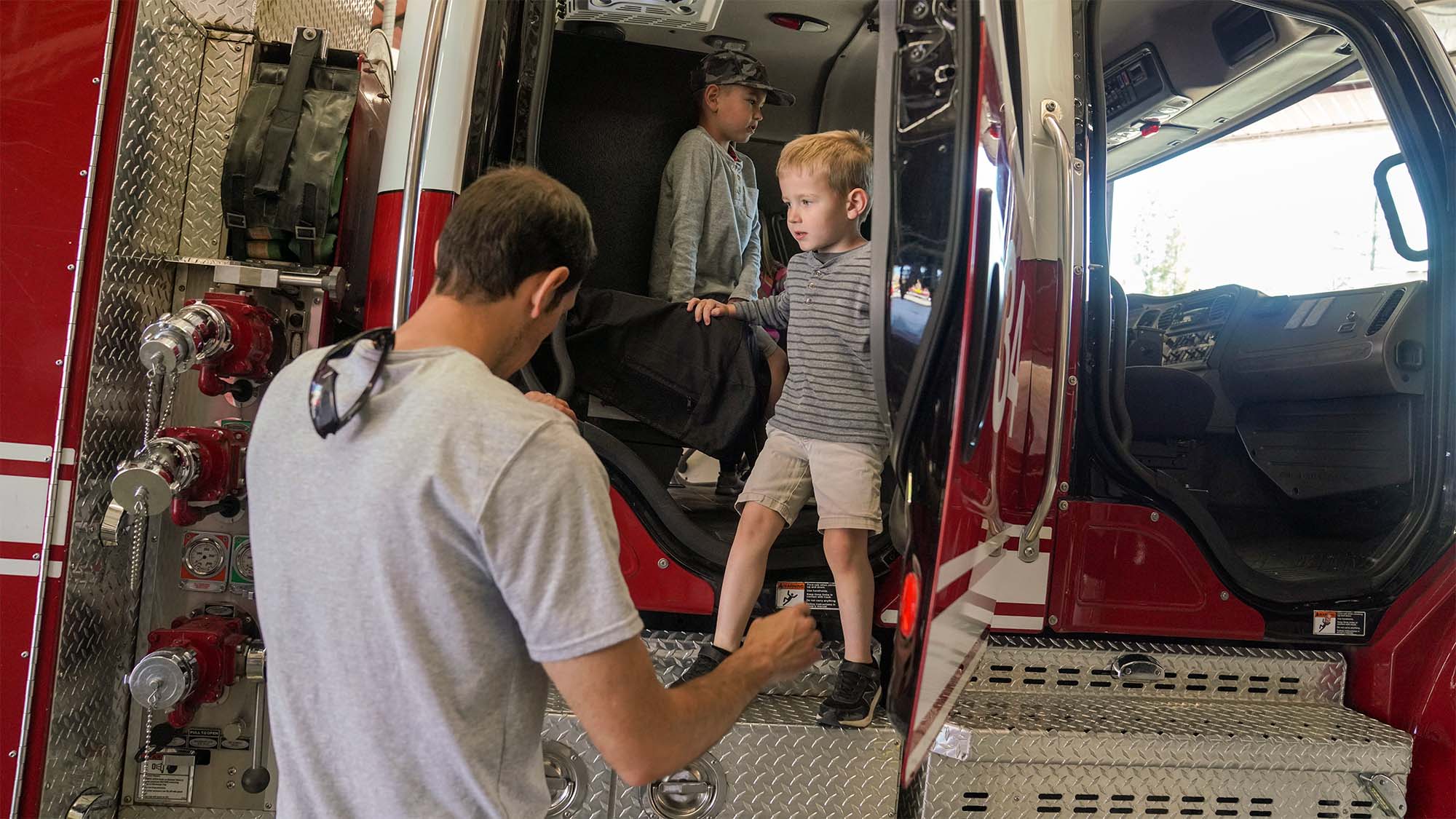 Adult helping two young boys climb into a red fire truck, photo