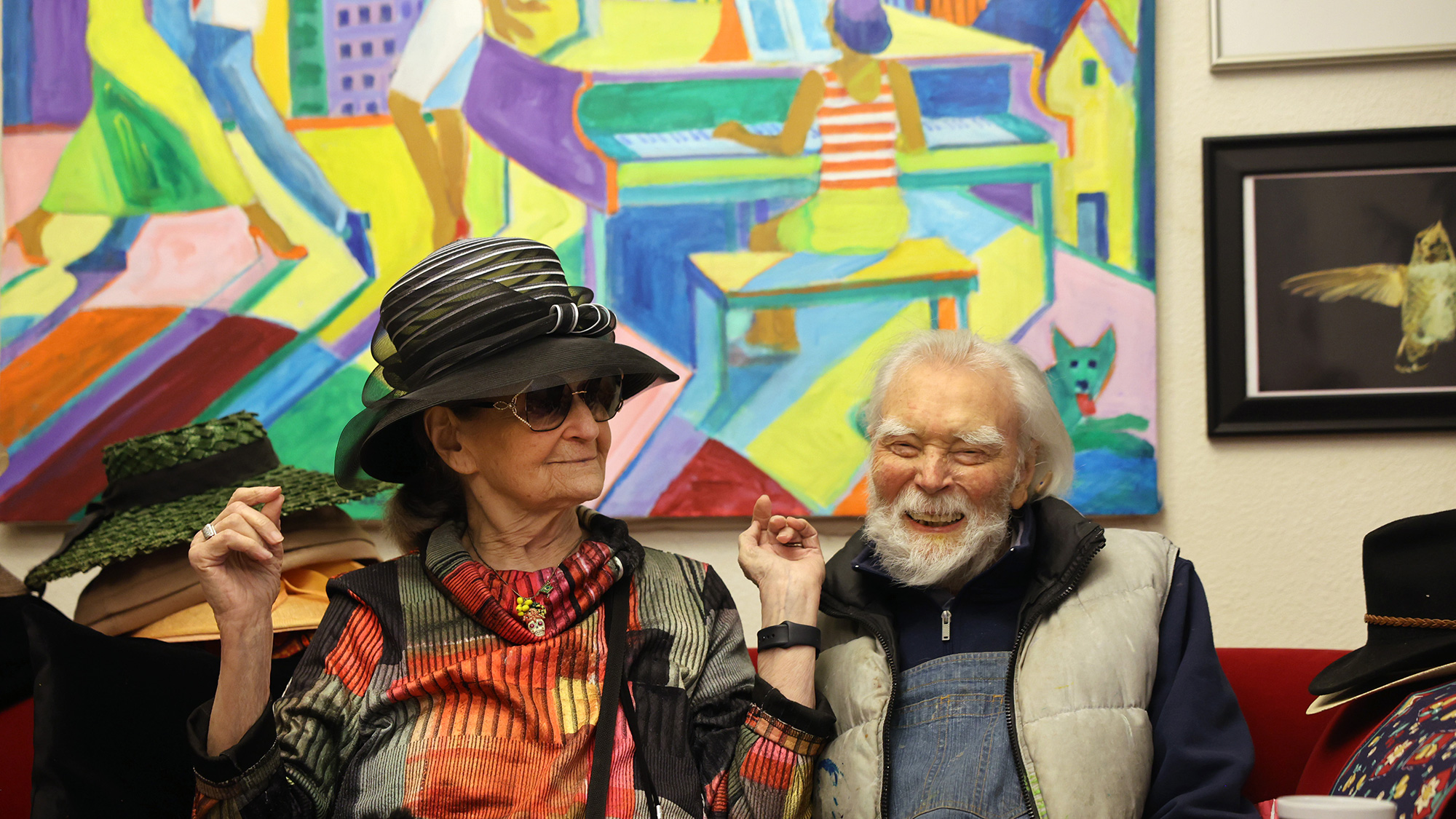 A man and woman with distinctive hats sit in front of a colorful painting, smiling.