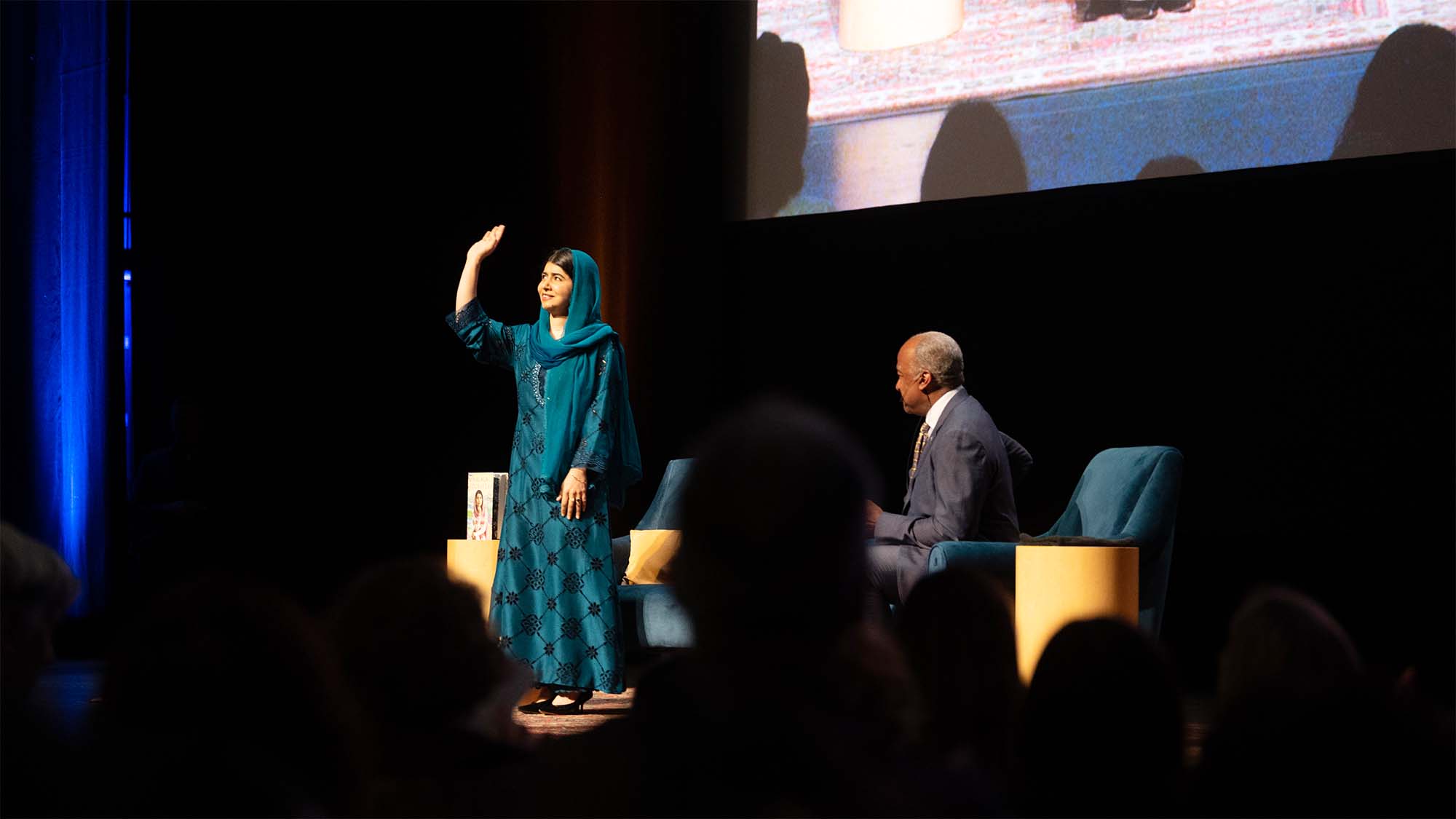 Nobel laureate Malala Yousafzai stands and waves to the crowd while on stage with Chancellor Gary S. May