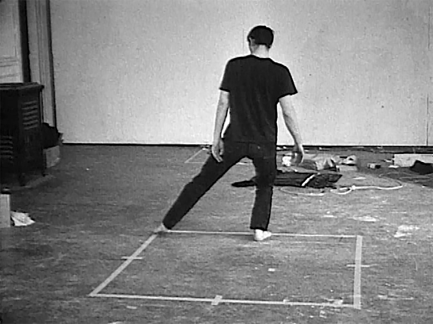 Black-and-white photo of person standing in taped square on scuffed studio floor, back turned. Bruce Nauman, Dance or Exercise on the Perimeter of a Square (Square Dance), 1967–68. Black-and-white video with sound, transferred from 16 mm film, 8:24 min. (Electronic Arts Intermix (EAI), New York. © 2026 Bruce Nauman / Artists Rights Society (ARS), New York)