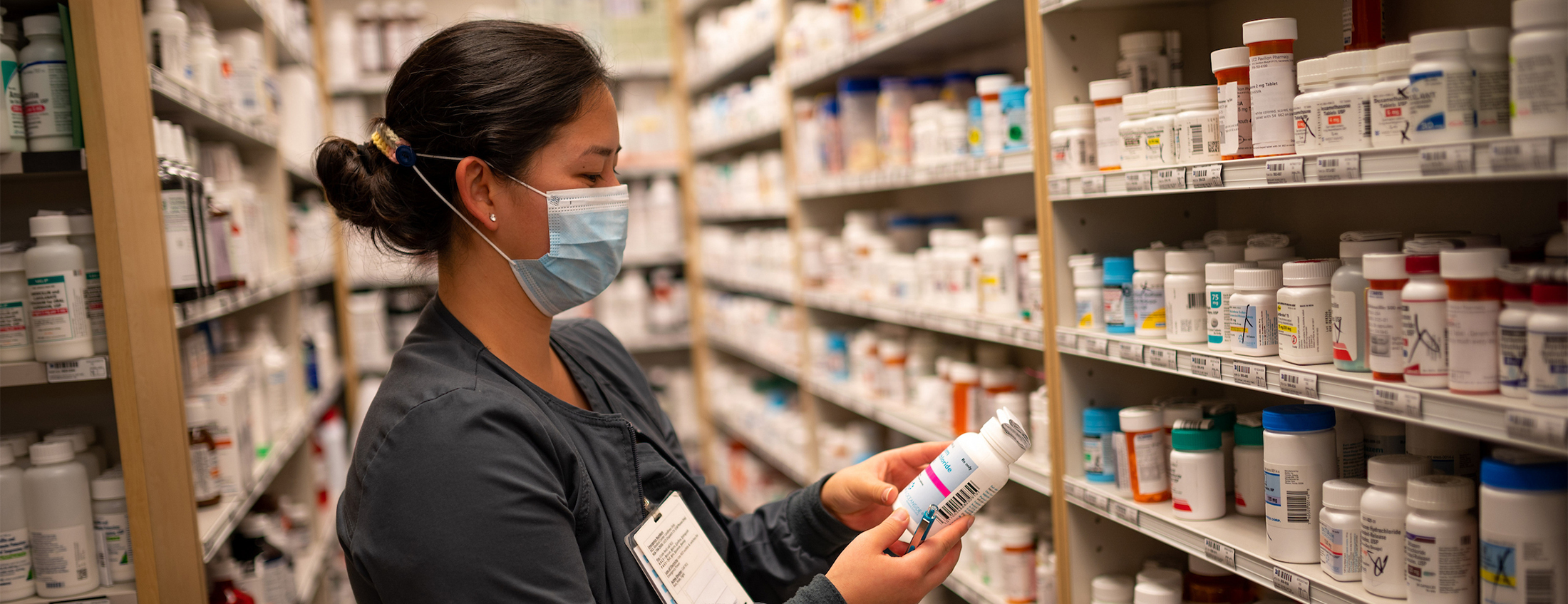 A pharmacist in a blue face mask stands between rows of shelving holding prescription medication and examines one of the bottles.