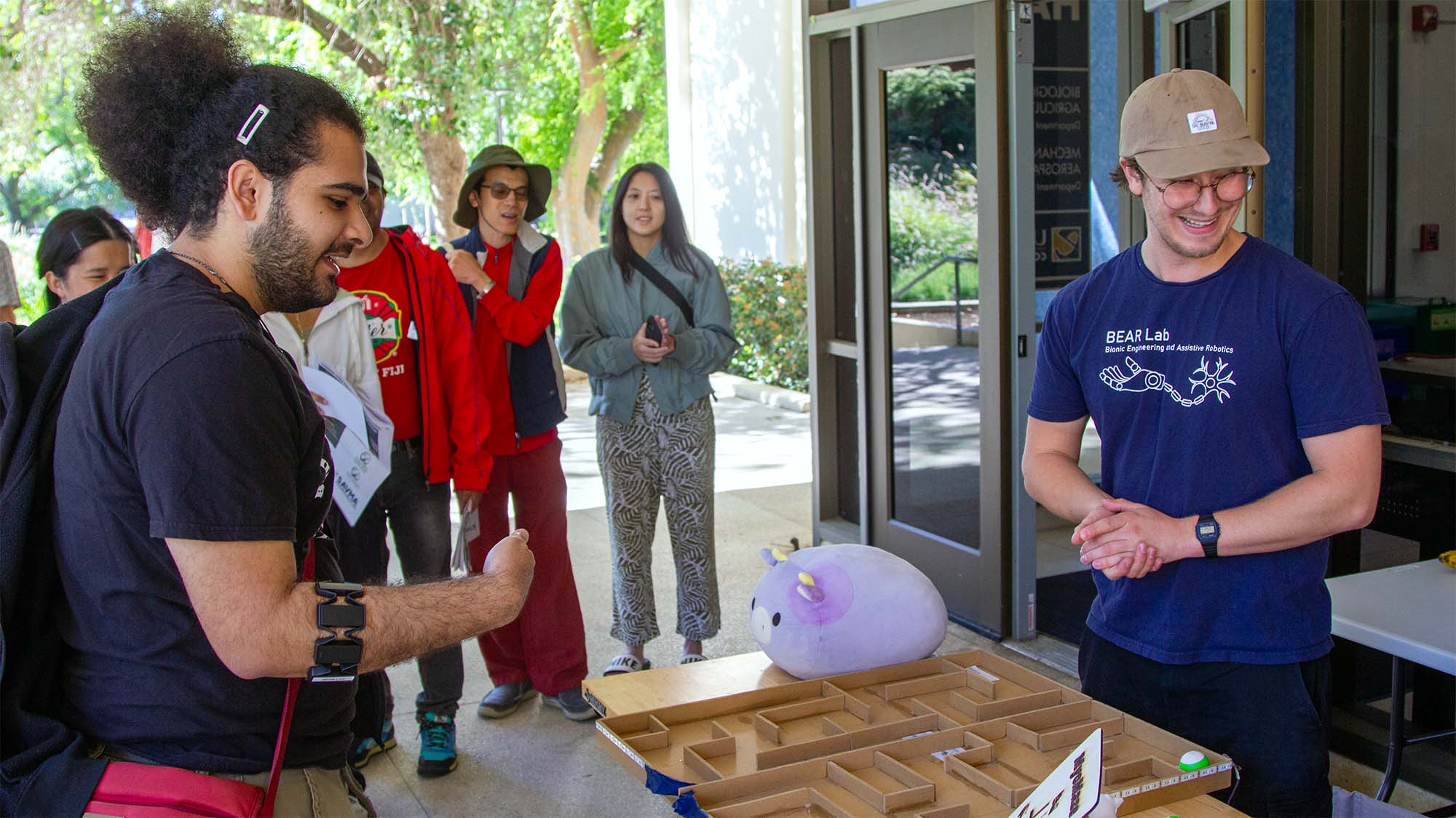 People at a tech demo, smiling presenter in blue shirt and cap by a wooden table