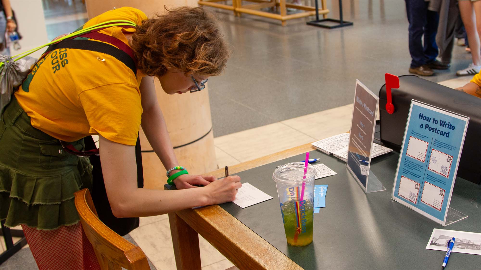 person in yellow shirt leans over wooden bench, writing at an event table