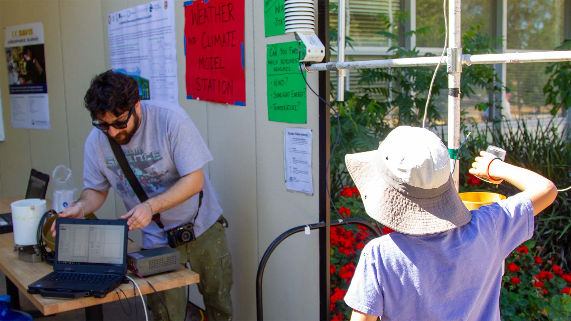 Man at table with laptop talks to child in sun hat by window with hanging plants.