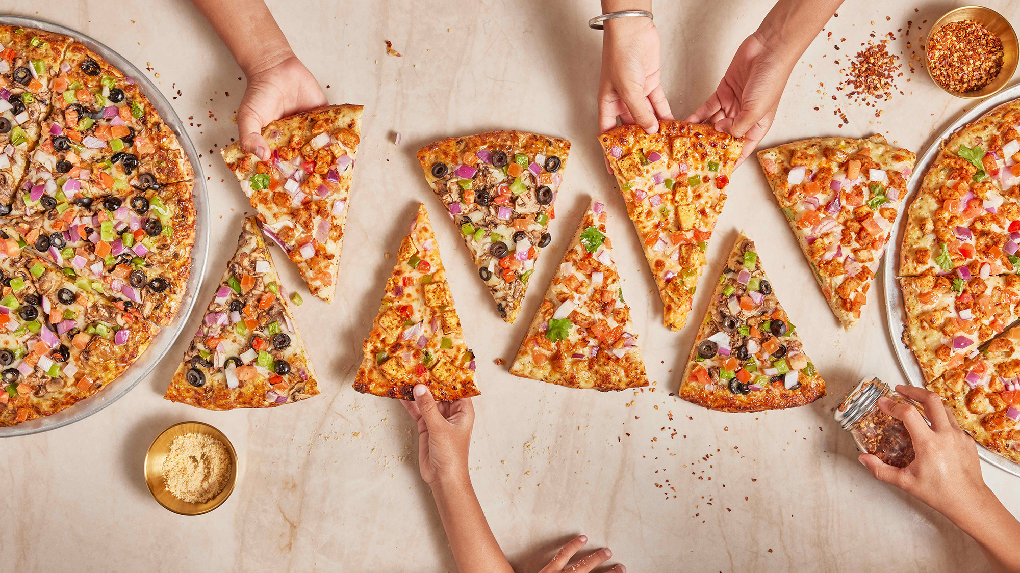 Hands reaching for slices of colorful pizza on a table with two pizza trays.