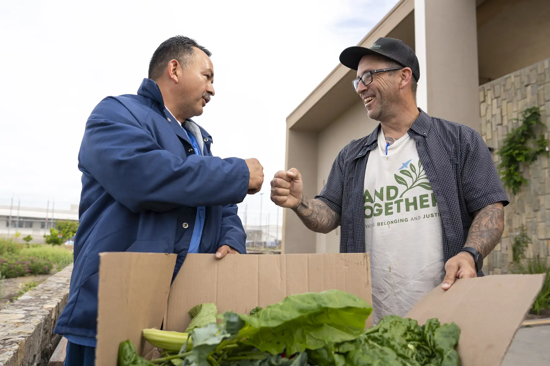 Two men smile and fist-bump while holding a box of fresh greens outdoors