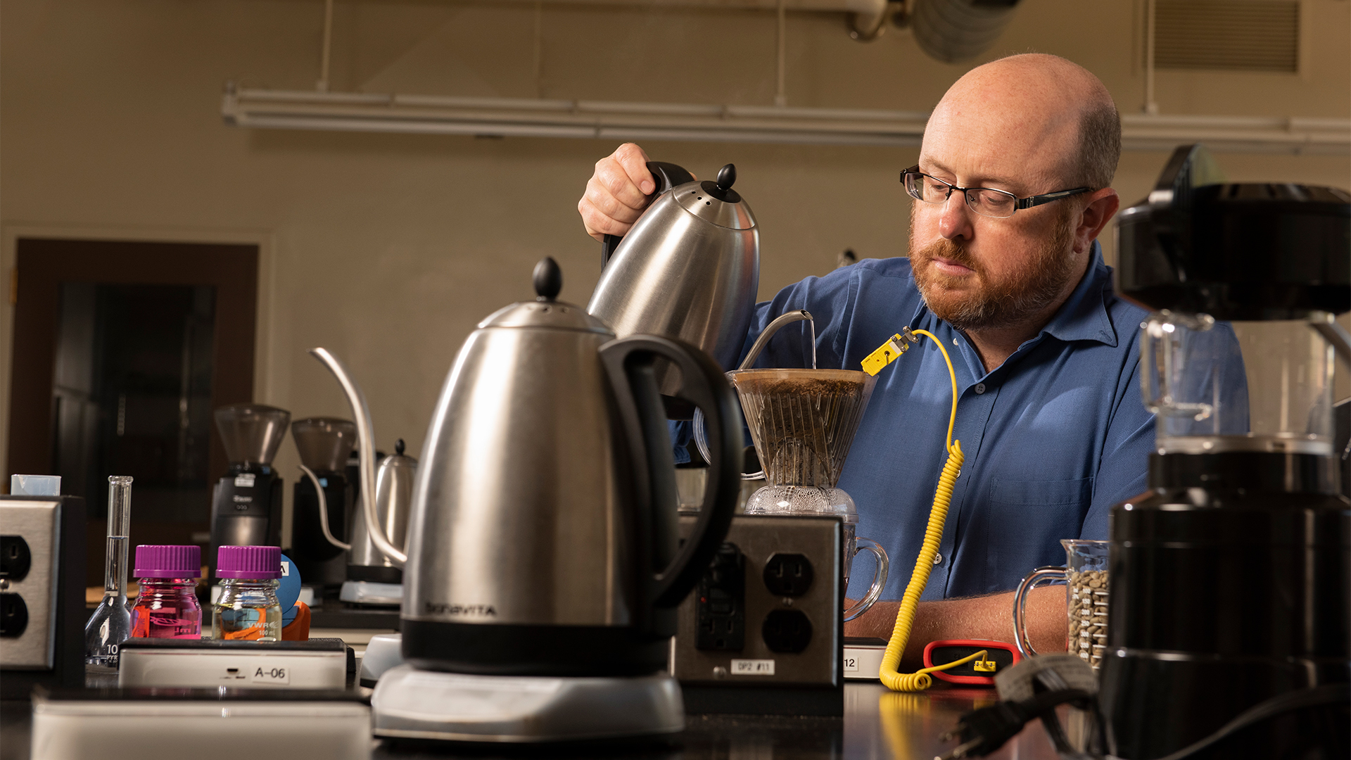 A bearded man in a blue button-down shirt and glasses concentrates while pouring hot water from a stainless-steel kettle over coffee grounds in a V60 plastic coffee dripper. He is surrounded by numerous scientific instruments and coffee tools.