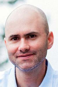 A smiling bald man with a short beard, wearing a light blue shirt, against a blurred background.