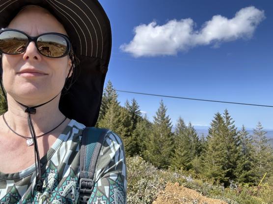 Light-complected woman in sunglasses in hat with landscape in background